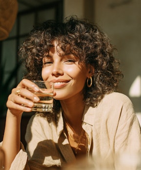 Femme aux cheveux bouclés souriante, un verre d’eau à la main, dans la lumière du soleil